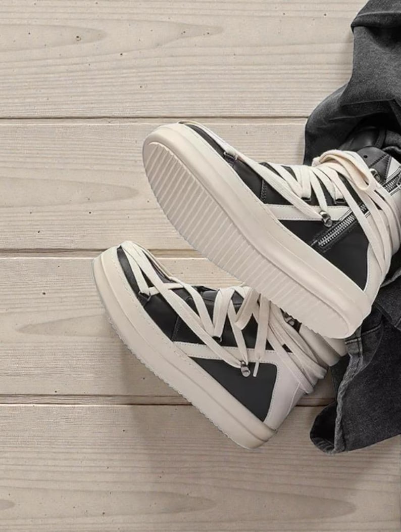 Pode incluir: T&eacute;nis de cano alto em preto e branco. Os sapatos apresentam uma parte superior em couro preto, atacadores brancos e uma sola grossa branca. Um detalhe de z&iacute;per &eacute; vis&iacute;vel na lateral. Colocados sobre uma superf&iacute;cie de madeira clara.