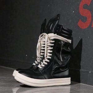 May include: Black high-top sneaker with a white sole and white laces. The sneaker has a zipper on the side and a wrap-around strap. The shoe is made of leather and has a unique design.