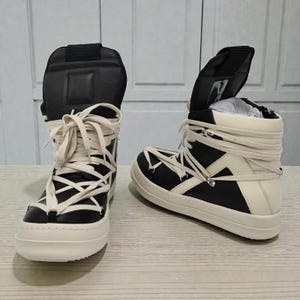 Pode incluir: Par de t&ecirc;nis de cano alto em preto e branco sujo. Os t&ecirc;nis apresentam um design exclusivo com cadar&ccedil;os creme cruzados e uma sola grossa branco sujo. A l&iacute;ngua &eacute; preta e a parte superior &eacute; uma mistura de pain&eacute;is pretos e branco sujo.