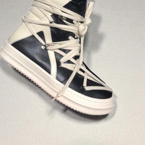 Pode incluir: T&ecirc;nis de cano alto em preto e creme. O t&ecirc;nis apresenta um design em camadas com uma sola grossa esbranqui&ccedil;ada e cadar&ccedil;os creme intrincados. A parte superior &eacute; uma combina&ccedil;&atilde;o de pain&eacute;is pretos e creme. A l&iacute;ngua &eacute; preta.