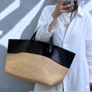 May include: A large tote bag with a woven straw base and a black leather top. The bag has two black leather handles and is held by a person wearing a white shirt.