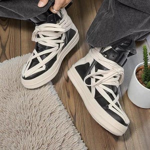 Pode incluir: T&ecirc;nis de cano alto preto e branco com cadar&ccedil;os creme enrolados na altura do tornozelo. Os t&ecirc;nis t&ecirc;m uma sola grossa, branca suja e um z&iacute;per na lateral. Os sapatos est&atilde;o em um piso de madeira.