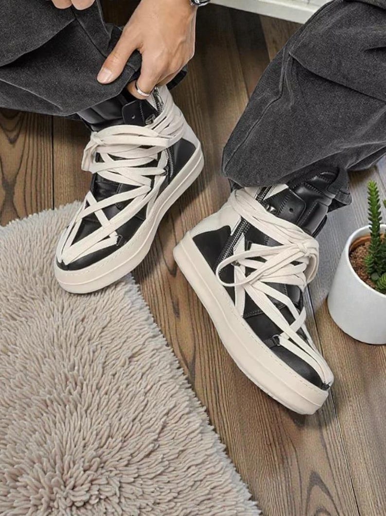 Pode incluir: T&ecirc;nis de cano alto preto e branco com cadar&ccedil;os creme enrolados na altura do tornozelo. Os t&ecirc;nis t&ecirc;m uma sola grossa, branca suja e um z&iacute;per na lateral. Os sapatos est&atilde;o em um piso de madeira.