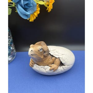 May include: A brown and tan dinosaur hatching from a white egg. The dinosaur has blue eyes and is emerging from a cracked eggshell. The sculpture is on a blue surface.