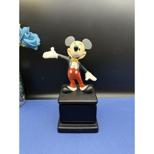 May include: A Mickey Mouse figurine in a classic pose, wearing a black tuxedo, red pants, and white gloves. The figurine stands on a black box-shaped base. The background is a dark blue.