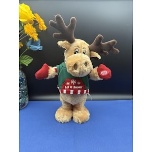 May include: A plush moose toy with brown antlers, a tan face, and a green sweater with the text "Let It Snow!". The moose wears red mittens and has a button on its right hand. The background is blue.