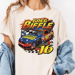 May include: Cream-coloured t-shirt with a colourful graphic of a race car, the text "Greg Biffle", and the number "16". The car design includes the words "National Guard" and a chequered flag pattern, with flames and stars.