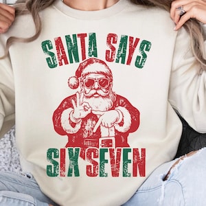 May include: Sand-colored sweatshirt with a graphic of Santa Claus wearing sunglasses and making a hand gesture. The text "SANTA SAYS SIX SEVEN" is printed above and below the Santa illustration in green and red.