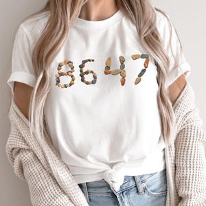 May include: White t-shirt with the number "8647" printed on the front. The numbers are made of small, colorful stones. The shirt is paired with blue jeans and a cream-colored cardigan.