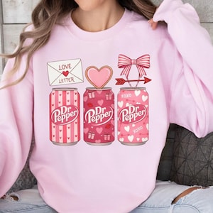 May include: Light pink sweatshirt featuring three Dr. Pepper soda cans with Valentine's Day themed designs. The cans have hearts, bows, and the text "XOXO". Above the cans are a love letter, a heart-shaped cookie, and a bow with an arrow.