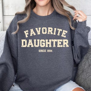 Puede incluir: Sudadera gris oscuro con el texto "FAVORITE DAUGHTER SINCE 1994" en una fuente desgastada de color blanquecino. La sudadera de cuello redondo est&aacute; hecha de un material suave. La imagen muestra la sudadera en una persona.