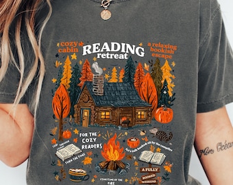 Op de afbeelding: Donkergrijs t-shirt met een kleurrijke afbeelding van een hut in een bos, pompoenen en een kampvuur. Tekst bevat "READING retreat", "cozy cabin" en "for the cozy readers". Het ontwerp heeft een herfstthema.