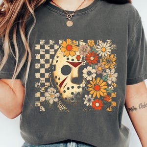 May include: Dark gray t-shirt featuring a graphic of a hockey mask with a floral design. The mask is yellow with red accents, set against a checkered background and a bouquet of colorful flowers. The shirt is a casual, vintage style.