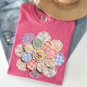 以下が含まれることがあります： 様々な柄の生地で作られた、大きなアップリケの花柄が特徴のピンク色のTシャツ。 花はシャツの中央に配置され、花柄、ギンガムチェック、無地の生地が組み合わされています。 ライトウォッシュのジーンズとベージュの帽子と組み合わせています。