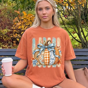 May include: A burnt orange short-sleeve t-shirt featuring a central graphic of a pumpkin with a large blue bow. The pumpkin design incorporates various patterns including leopard print, orange and white gingham, and blue floral motifs.