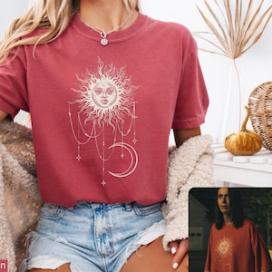 May include: A crimson short-sleeved t-shirt featuring a cream-colored graphic print of a sun with a face, a crescent moon, and hanging beaded chains with star accents. The shirt has a crew neckline and a relaxed fit, worn with light blue denim shorts.