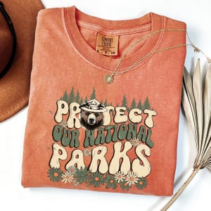 Puede incluir: Una camiseta naranja Comfort Colors con el texto "Protect Our National Parks" en una fuente retro, con un oso con un sombrero de guardabosques. El dise&ntilde;o incluye &aacute;rboles verdes y margaritas. Un collar dorado est&aacute; drapeado sobre la camisa.