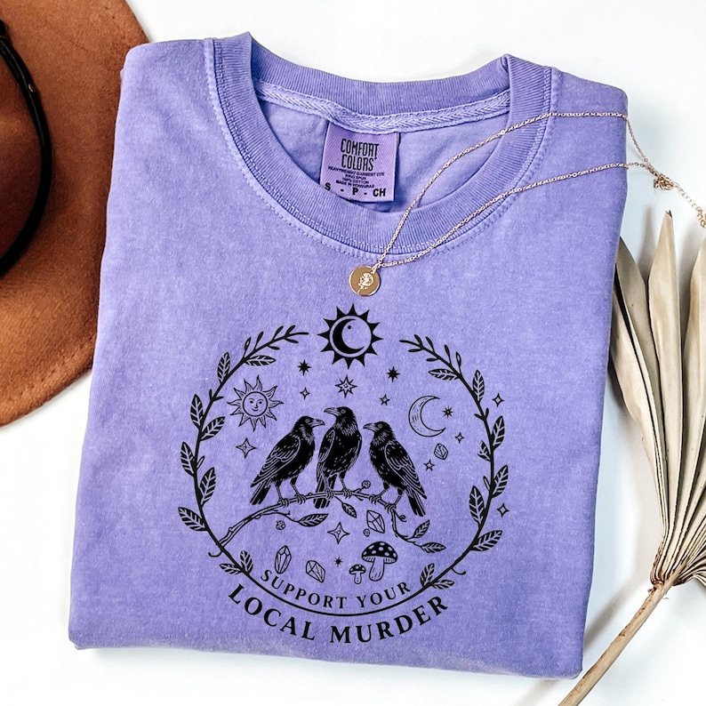 May include: A light purple Comfort Colors t-shirt with a black graphic of three crows perched on a branch, surrounded by celestial and botanical elements. The text "Support Your Local Murder" is printed below the design. A gold necklace is draped across the shirt.