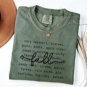 K&ouml;nnte beinhalten: Olivgr&uuml;nes Comfort Colors T-Shirt mit dem Wort "fall" in Schreibschrift und einer Liste von W&ouml;rtern zum Thema Herbst. Eine goldene Halskette mit Anh&auml;nger ist &uuml;ber dem Shirt drapiert.