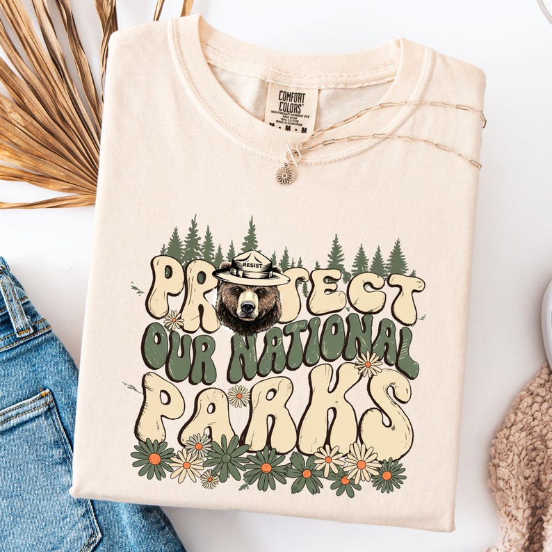 Puede incluir: Camiseta color crema con el texto "PROTECT OUR NATIONAL PARKS" en una fuente retro, con un oso con sombrero de guardabosques. El dise&ntilde;o incluye &aacute;rboles verdes y detalles de margaritas, con la palabra "RESIST" en el sombrero.