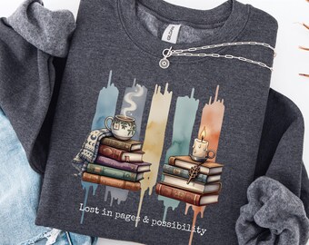 May include: Dark heather grey sweatshirt featuring a graphic of books, a teacup, and a candle. The text "Lost in pages & possibility" is printed below the graphic. A silver chain necklace with a sun pendant is also visible.