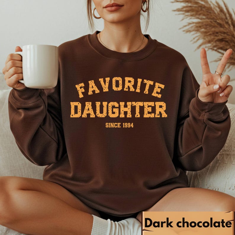 Puede incluir: Sudadera marr&oacute;n chocolate oscuro con el texto "FAVORITE DAUGHTER SINCE 1994" en una fuente naranja desgastada. La sudadera es de cuello redondo y la lleva puesta una persona.
