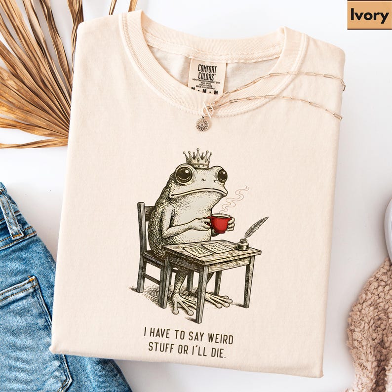 K&ouml;nnte beinhalten: Elfenbeinfarbenes Comfort Colors T-Shirt mit einer Illustration einer gekr&ouml;nten Froschfigur, die eine rote Tasse h&auml;lt und an einem Schreibtisch sitzt. Der Text "I HAVE TO SAY WEIRD STUFF OR I'LL DIE" ist darunter gedruckt. Eine Halskette liegt auf dem Shirt.