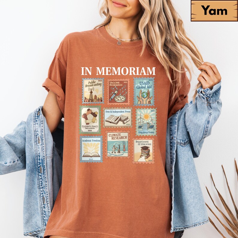 May include: Rust-colored t-shirt with the words "IN MEMORIAM" above a collection of postage stamp-style graphics. The stamps depict various themes, including "Public Broadcasting," "Vaccine Research," and "USAID Global Aid."