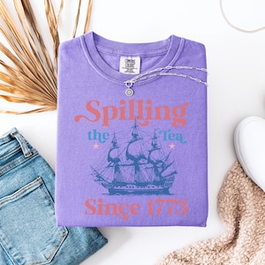 May include: Purple t-shirt with a vintage-style graphic of a ship and the text "Spilling the Tea Since 1773".