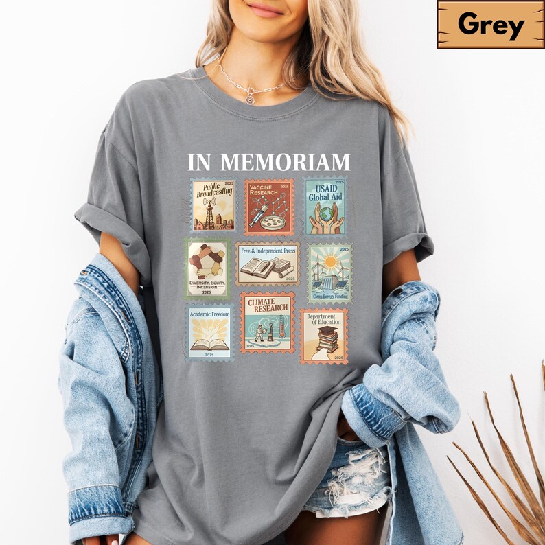 May include: Grey t-shirt with the words "IN MEMORIAM" above nine postage stamp-style graphics. The graphics include text such as "Public Broadcasting", "Vaccine Research", and "Climate Research". The shirt is a casual style.