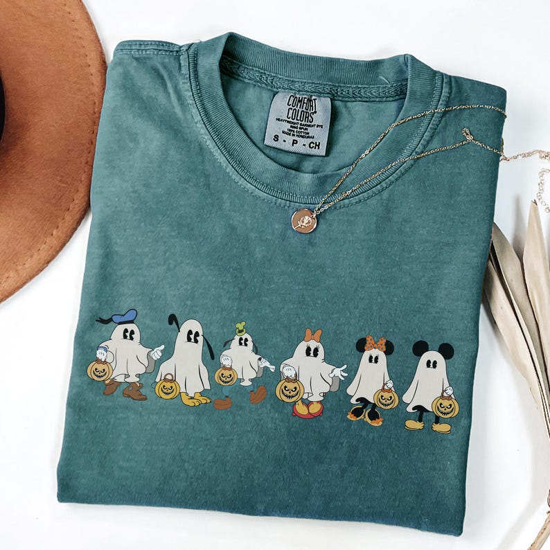 Op de afbeelding: Teal Comfort Colors T-shirt met een Halloween-thema van cartoongeesten met pompoenen. Het shirt heeft een ronde hals en korte mouwen. Een gouden ketting met een hanger is over het shirt gedrapeerd.