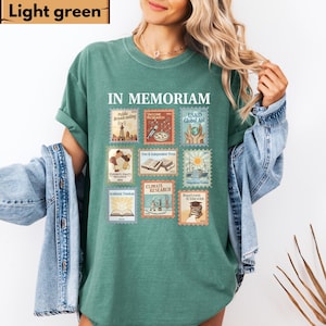 May include: Light green t-shirt with the words "IN MEMORIAM" and postage stamp-like graphics. The stamps depict themes like public broadcasting, vaccine research, and climate research. The shirt is a solid light green color.