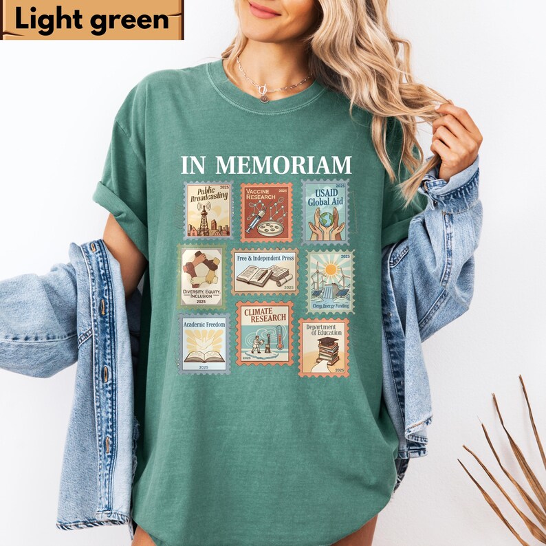 May include: Light green t-shirt with the words "IN MEMORIAM" and postage stamp-like graphics. The stamps depict themes like public broadcasting, vaccine research, and climate research. The shirt is a solid light green color.