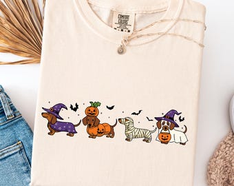 May include: Ivory-colored sweatshirt featuring a Halloween-themed graphic of dachshunds in costumes: a witch, a pumpkin, a mummy, and a ghost. The Comfort Colors shirt has a crew neck and long sleeves. A gold necklace with a sun pendant is also visible.