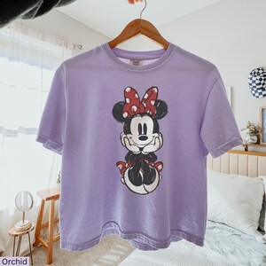 May include: Orchid-colored t-shirt featuring a Minnie Mouse graphic. The graphic shows Minnie Mouse with a red polka-dot bow, black ears, and a smiling face. The shirt is hanging on a wooden hanger.