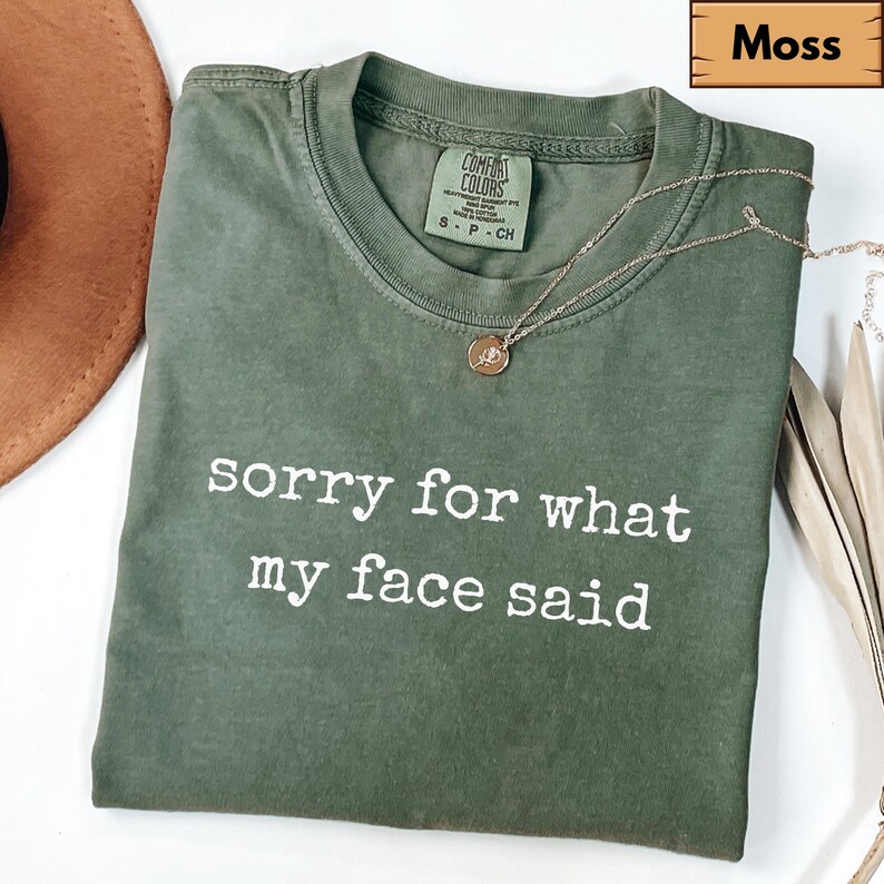 May include: A moss green t-shirt with the text "sorry for what my face said" in white. The shirt has a crew neck and short sleeves. A gold necklace with a pendant is draped across the shirt.