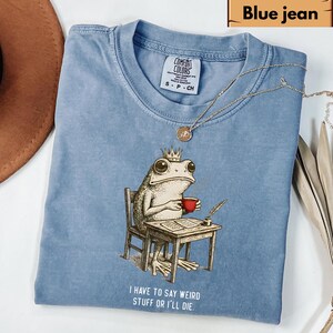 K&ouml;nnte beinhalten: Blaues Jeans-T-Shirt mit einer Frosch-Illustration. Der Frosch tr&auml;gt eine Krone und h&auml;lt eine rote Tasse, sitzt an einem Schreibtisch mit Feder und Tinte. Der Text auf dem Shirt lautet: "I HAVE TO SAY WEIRD STUFF OR I'LL DIE."