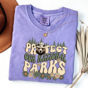 Puede incluir: Una camiseta lila Comfort Colors con el texto "PROTECT OUR NATIONAL PARKS" en una fuente retro, con un oso con sombrero de guardabosques. El dise&ntilde;o incluye &aacute;rboles y flores. Un collar dorado est&aacute; drapeado sobre la camiseta.
