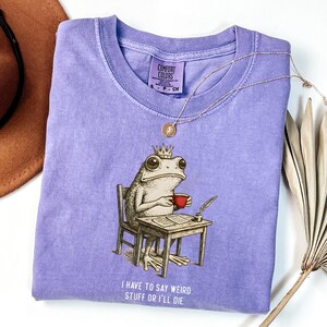 K&ouml;nnte beinhalten: Ein lila Comfort Colors T-Shirt mit einer skurrilen Illustration einer Froschk&ouml;nigin, die an einem Schreibtisch sitzt, eine rote Tasse h&auml;lt und mit einer Feder schreibt. Der Text "I HAVE TO SAY WEIRD STUFF OR I'LL DIE" ist darunter gedruckt. Eine goldene Halskette liegt auf dem Shirt.