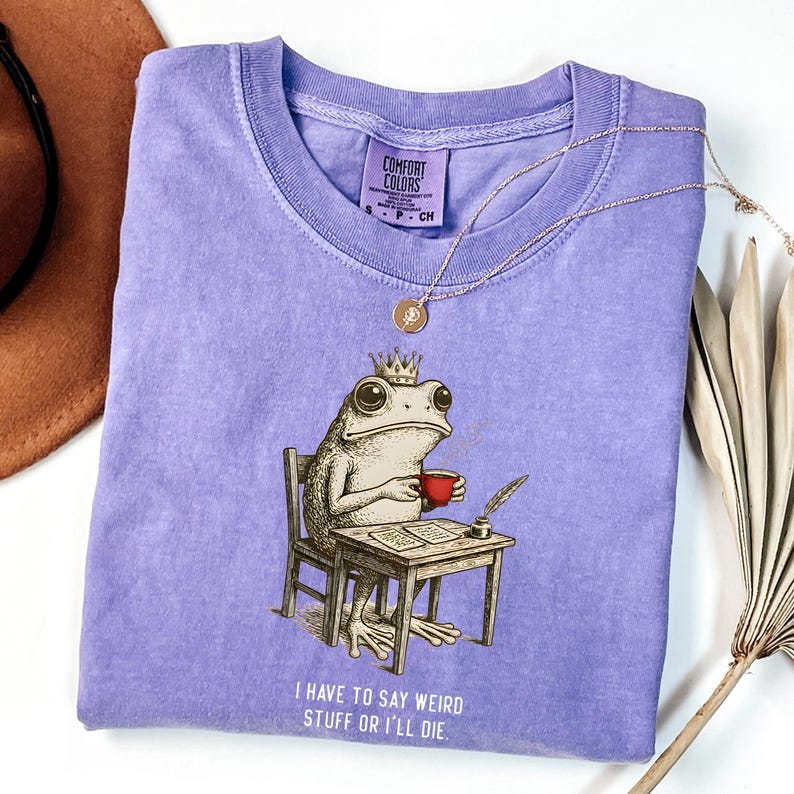 K&ouml;nnte beinhalten: Ein lila Comfort Colors T-Shirt mit einer skurrilen Illustration einer Froschk&ouml;nigin, die an einem Schreibtisch sitzt, eine rote Tasse h&auml;lt und mit einer Feder schreibt. Der Text "I HAVE TO SAY WEIRD STUFF OR I'LL DIE" ist darunter gedruckt. Eine goldene Halskette liegt auf dem Shirt.