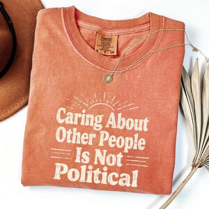 Puede incluir: Una camiseta de color coral con el texto "Caring About Other People Is Not Political" en blanco, con una fuente de estilo vintage. Tambi&eacute;n se ve un collar dorado con un colgante y un sombrero marr&oacute;n.