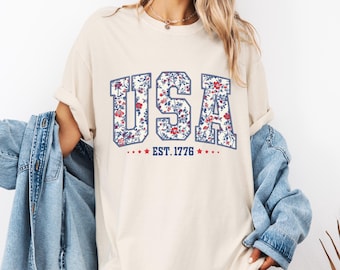 May include: Cream-colored t-shirt with the word "USA" in a floral pattern, outlined in blue. Below the text is "EST. 1776" with red stars. The shirt is worn with a denim jacket.