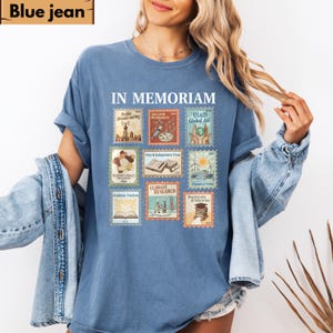 May include: A blue jean colored t-shirt with the words "IN MEMORIAM" at the top. The shirt features nine postage stamp-style graphics with text including "Public Broadcasting", "Vaccine Research", and "USAID Global Aid".
