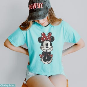 May include: A light blue t-shirt featuring a Minnie Mouse graphic. The cartoon character is depicted with a red bow with white polka dots. The shirt is a solid color and has short sleeves.