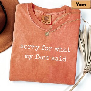 May include: A yam-colored t-shirt with the text "sorry for what my face said" in white. A gold necklace with a pendant rests on the shirt. The shirt is folded and has a Comfort Colors tag.