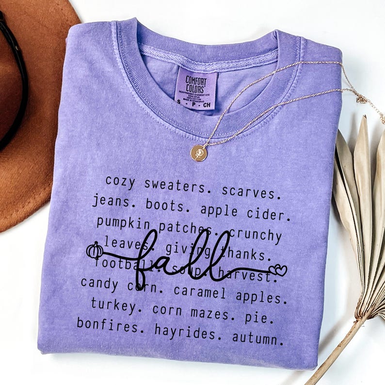 K&ouml;nnte beinhalten: Ein lila Comfort Colors T-Shirt mit dem Wort "fall" in Schreibschrift, umgeben von einer Liste von W&ouml;rtern zum Thema Herbst. Eine goldene Halskette mit Anh&auml;nger und ein brauner Hut sind ebenfalls zu sehen.