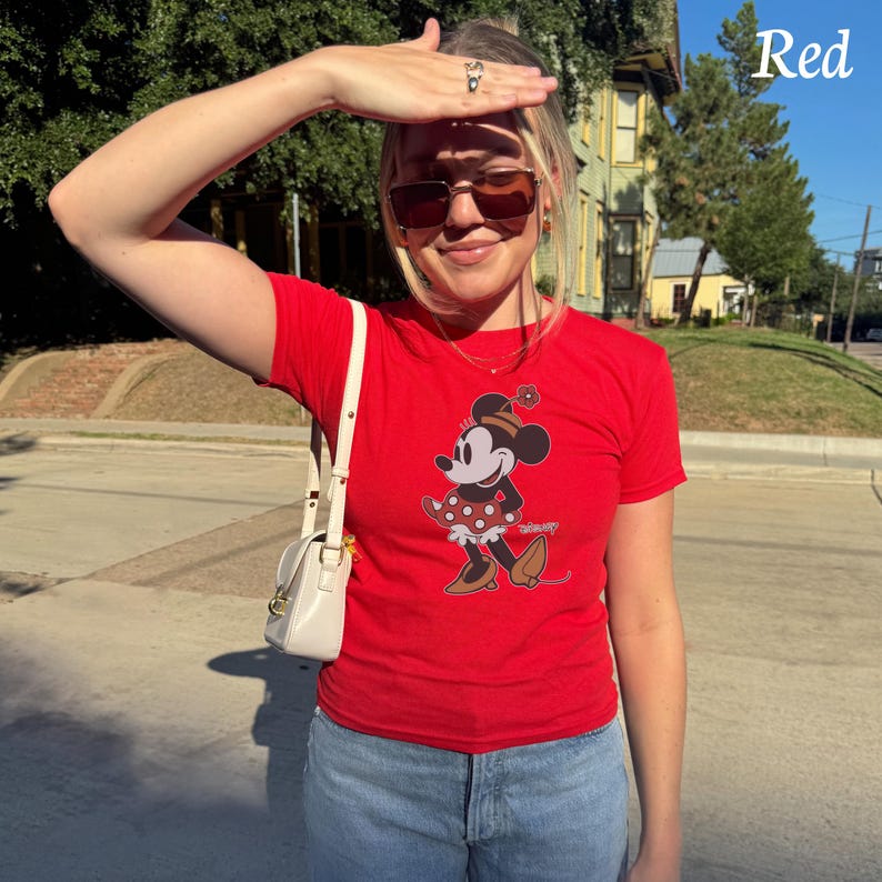 May include: A red t-shirt featuring a Minnie Mouse graphic. The cartoon character is depicted in a polka-dot dress and hat. The word "Red" is in the upper right corner. The person is wearing sunglasses and a white purse.