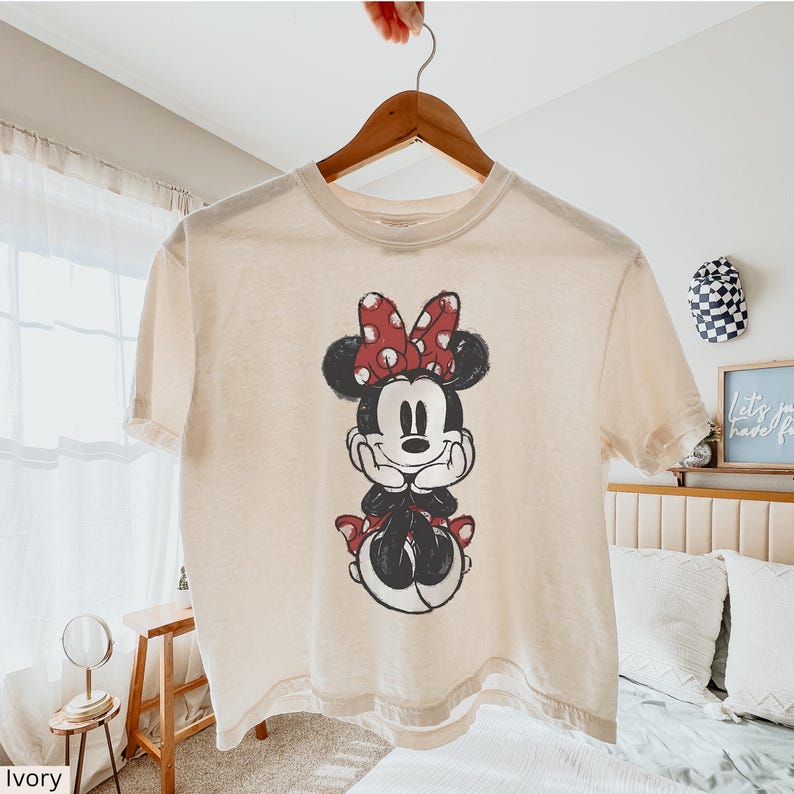 May include: Ivory-colored t-shirt featuring a graphic of Minnie Mouse with a red polka-dot bow. The cartoon character is depicted in black and white with a red bow and dress accents. The shirt is hanging on a wooden hanger.