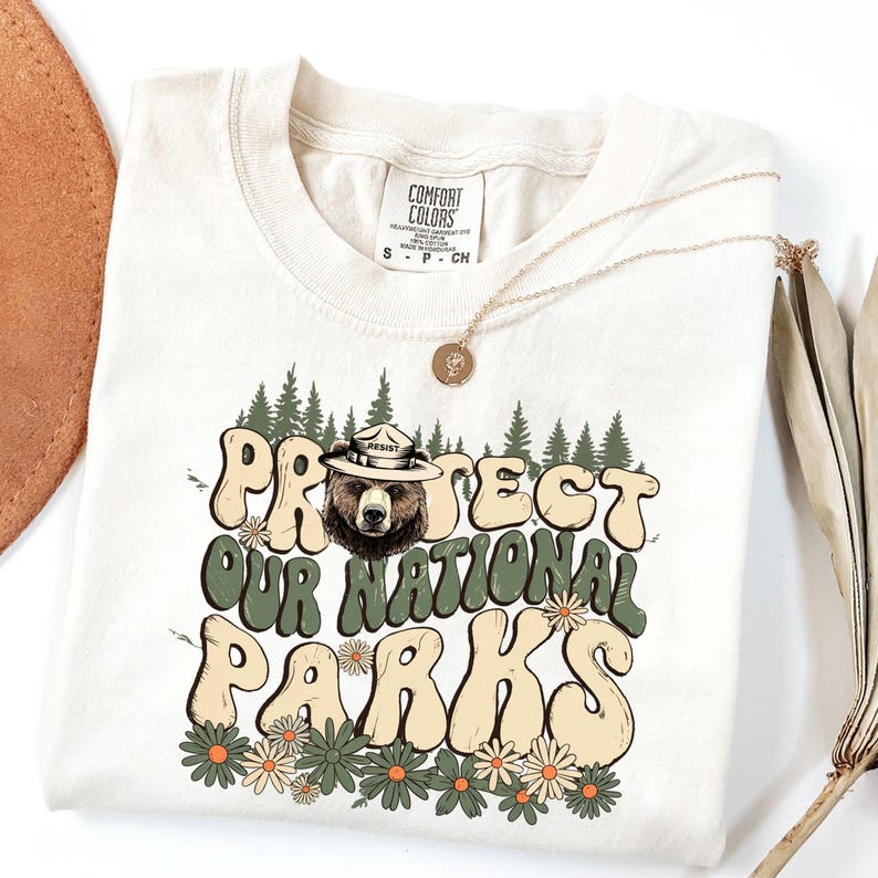 Puede incluir: Camiseta color crema con el texto "PROTECT OUR NATIONAL PARKS" en una fuente retro, con un oso que lleva un sombrero de guardabosques. El dise&ntilde;o incluye &aacute;rboles y flores verdes. Un collar de oro est&aacute; drapeado sobre la camiseta.