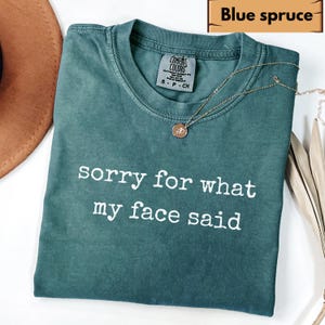 May include: Blue spruce Comfort Colors t-shirt with the text "sorry for what my face said" in white. A gold necklace with a pendant is draped across the shirt. The shirt is folded.